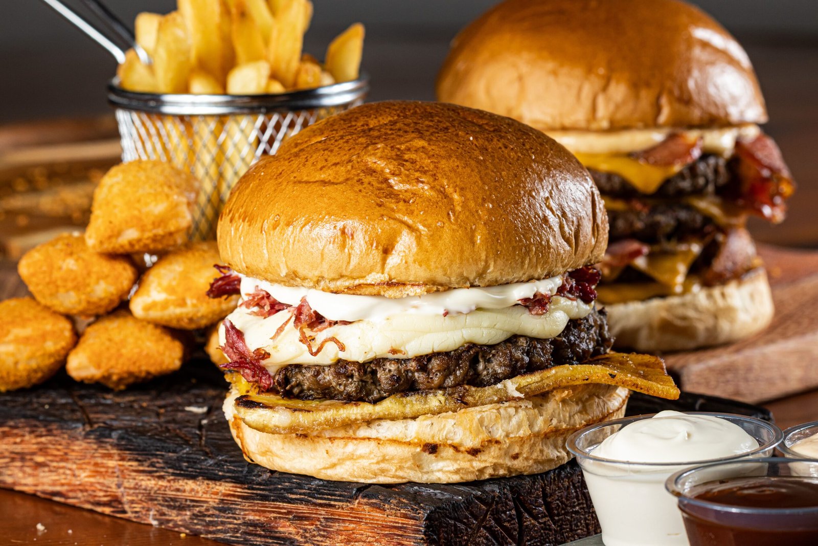 Delicious gourmet burgers with cheese, fries, and dipping sauces on a rustic board.